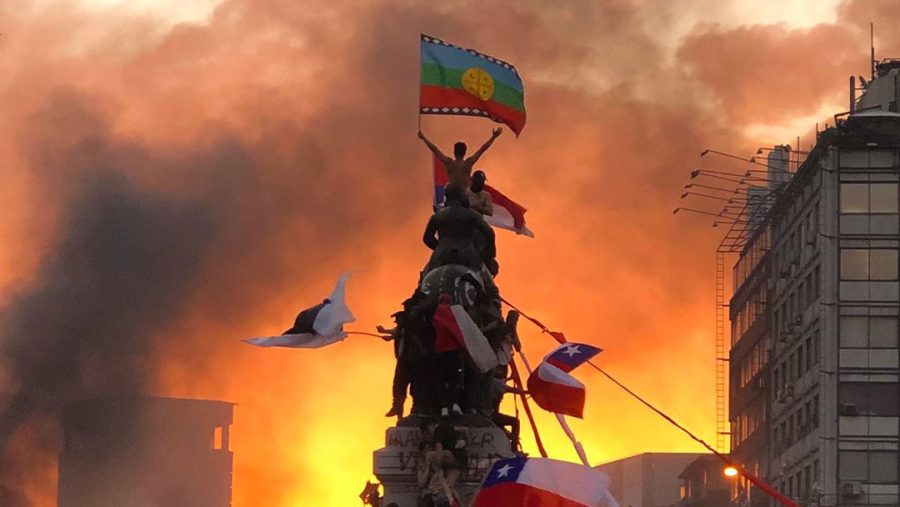 Protester+stands+at+the+top+of+a+military+statue+waving+the+Mapuche+flag%2C+an+indeginous+group+known+for+their+resistance%2C+in+Santiago%2C+Chile.