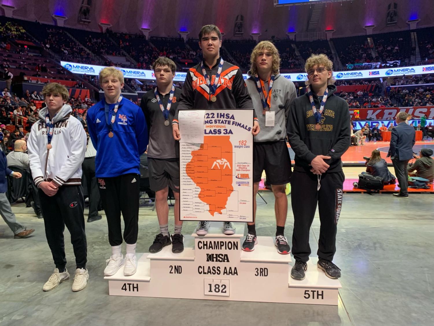 Knudten stands on the first position place on the podium after his win in the competition. He holds a “2022 IHSA State Finals Class 3A” poster. He also wears a medal that declares him as the IHSA 3A 182 lb State Champion.