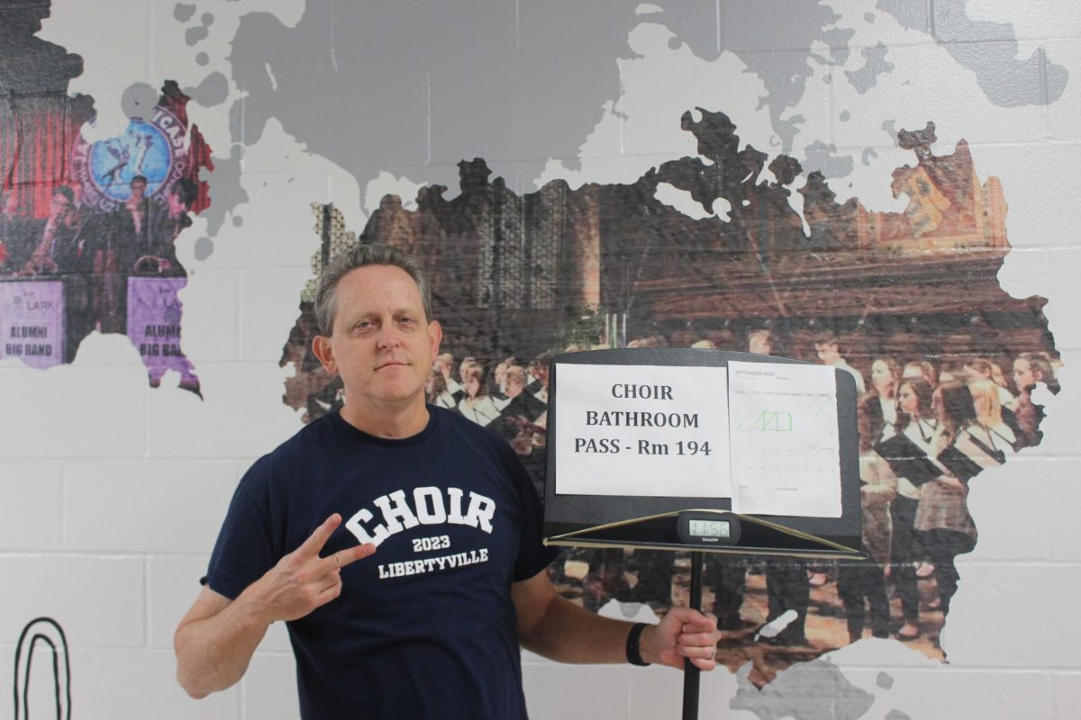 Choir teacher Dr. Brown poses with his student-made music stand hall pass. When Dr. Brown needed to create a hall pass, he sought out the opinion of his senior students for ideas. “I think in the whole school [teachers] were all trying to come up with our own bathroom pass scenario,” Dr. Brown said. 