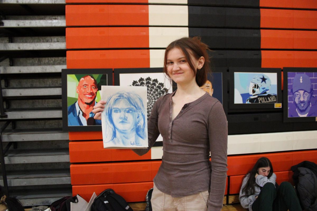 Senior Ava Sieberlich holds up her portrait drawn for her by sophomore Angela Xia during the art show. Each figure drawing was made within ten minutes with chalk pastels on paper.